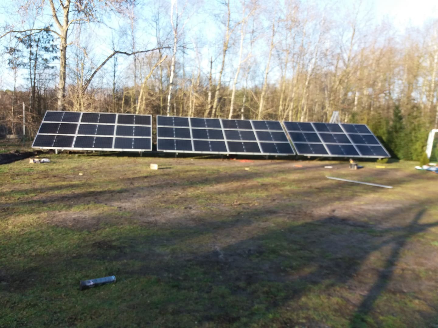 Trzy sekcje paneli słonecznych na metalowym stelażu, rozstawione na trawiastym terenie z drzewami w tle w słoneczny dzień.