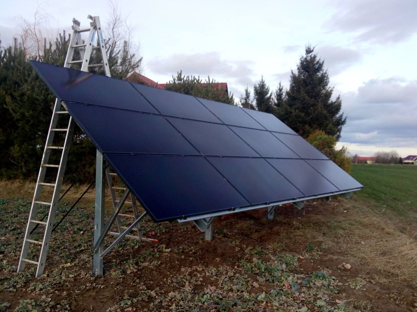 Czarny panel słoneczny na metalowej konstrukcji, z aluminiową drabiną opartą o jego górną krawędź na tle pochmurnego nieba i zieleni.
