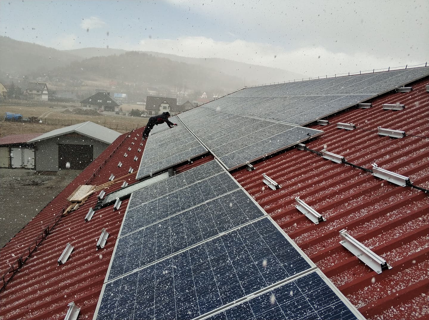 Instalacja paneli słonecznych na czerwonym dachu podczas opadów śniegu, widoczny pracownik w czarnym kombinezonie oraz górzysty krajobraz w tle.