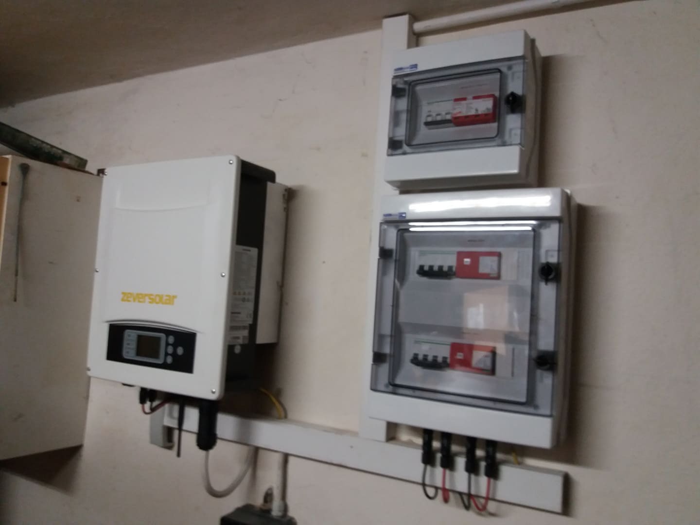 Biały inwerter marki Zeversolar zamontowany na ścianie obok dwóch białych skrzynek z zabezpieczeniami elektrycznymi, widoczne przewody i rury instalacyjne.