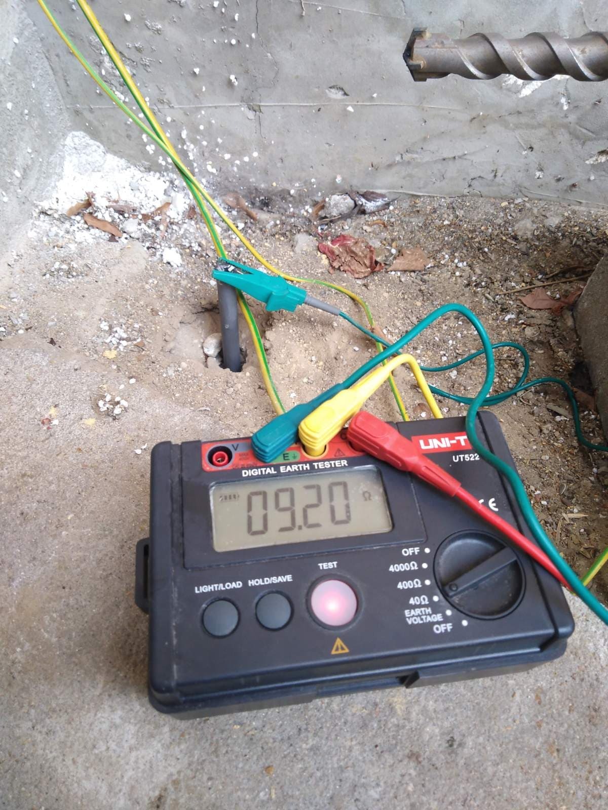 Pomiar rezystancji uziemienia za pomocą miernika UNI-T UT522, widoczne przewody pomiarowe z krokodylkami podłączone do uziomu w gruncie. Na wyświetlaczu miernika wartość 09.20 omów. W tle fragment...
