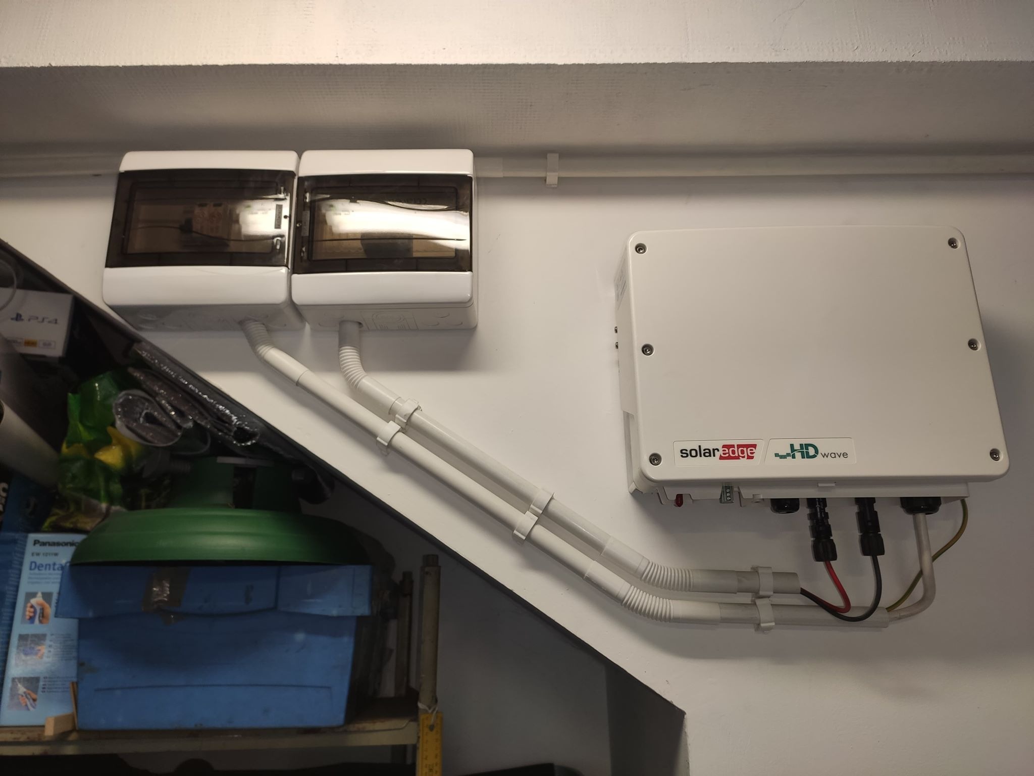 Inwerter SolarEdge HD-Wave zamontowany na białej ścianie, z widocznymi przewodami i dwoma skrzynkami bezpiecznikowymi powyżej, w tle półka z narzędziami i pudełkami.
