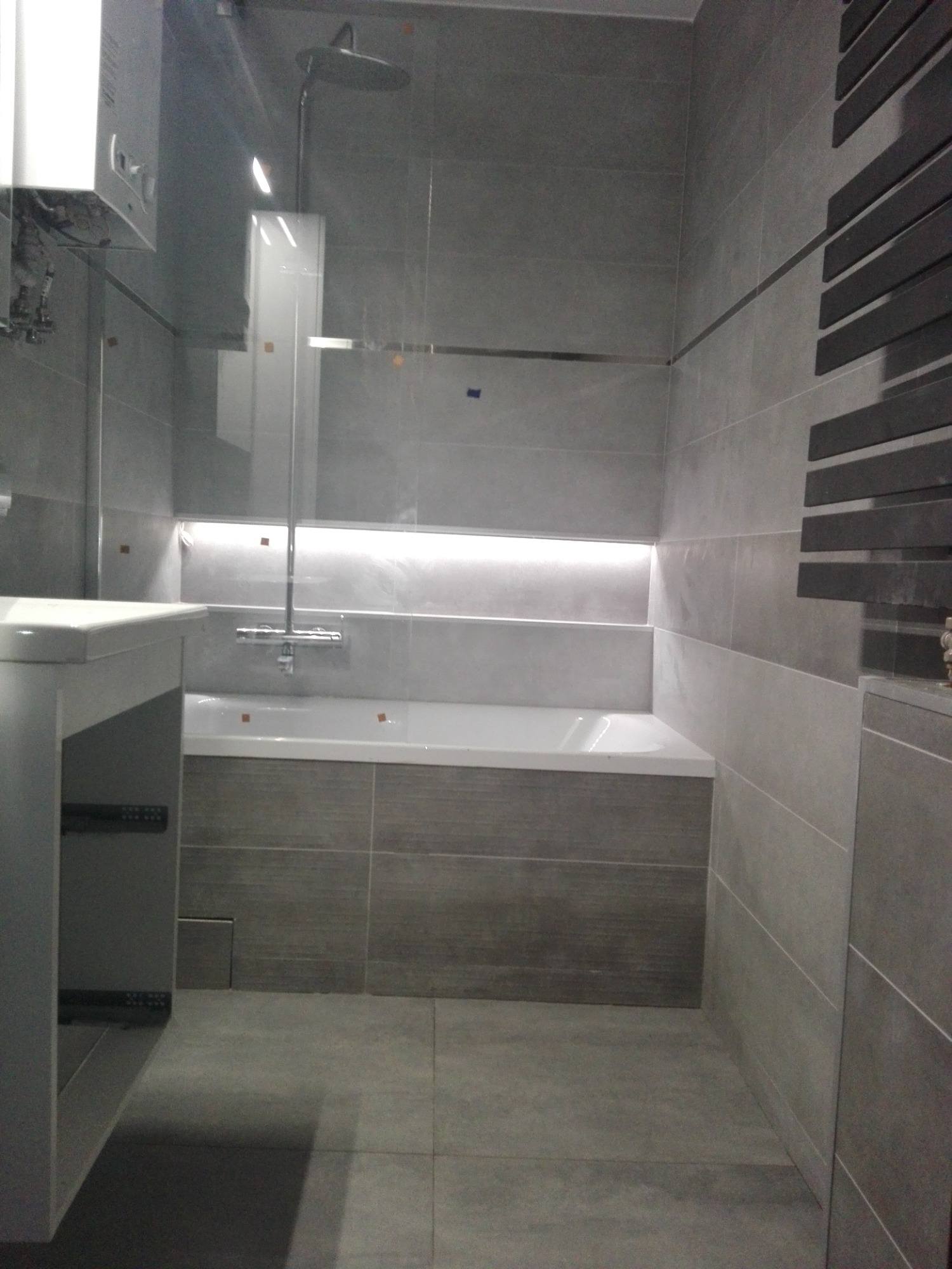 Nowoczesna łazienka w odcieniach szarości z podświetlaną wnęką nad wanną i grzejnikiem drabinkowym.