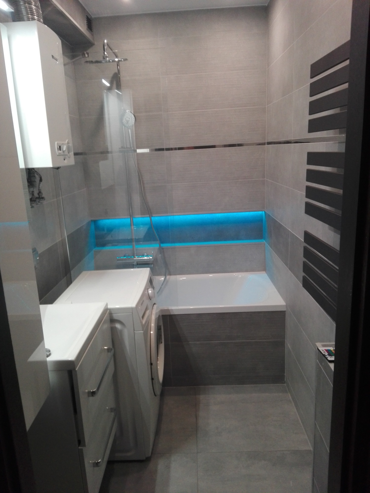 Nowoczesna łazienka z szarymi płytkami, podświetlaną wnęką nad wanną, grzejnikiem drabinkowym i pralką obok umywalki z szafką.