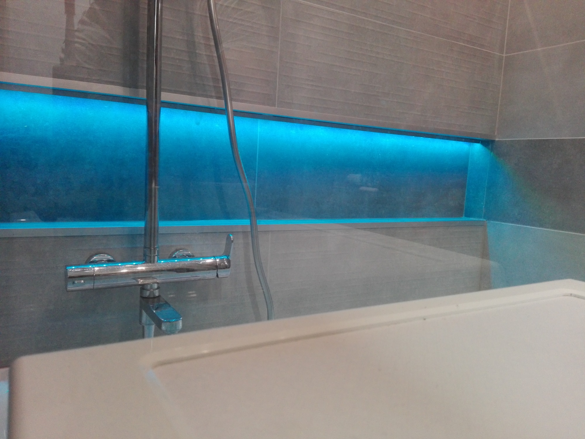 Szczegółowe ujęcie chromowanej baterii wannowej na tle szarej, podświetlanej wnęki w płytkach.