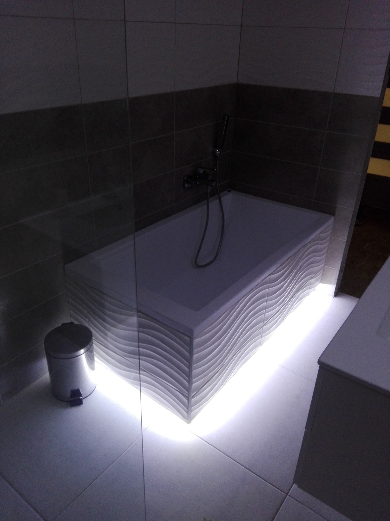 Nowoczesna wanna z falistym panelem przednim i podświetleniem LED w szaro-białej łazience z koszem na śmieci w rogu.