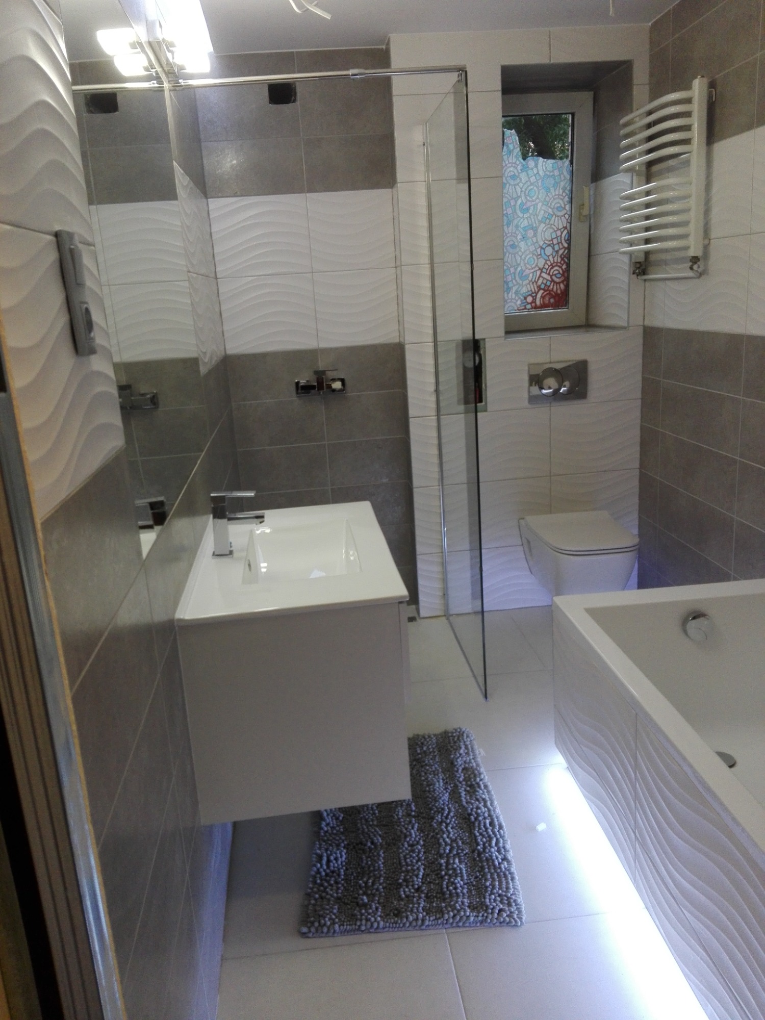 Nowoczesna łazienka z szaro-białymi płytkami, podświetlaną wanną i umywalką na szafce, z szarym dywanikiem i oknem z dekoracyjną folią.