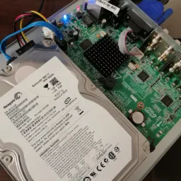 Wnętrze rejestratora monitoringu z podłączonym dyskiem twardym Seagate Barracuda 1TB, widoczna płyta główna z elementami elektronicznymi, kable SATA i zasilające, złącza BNC i Ethernet.