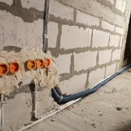 aWIĘC-ELEKTRO - Surowa ściana z betonu komórkowego w trakcie prac instalacyjnych, widoczne pomarańczowe puszki elektryczne zalane gipsem, peszel z przewodami oraz kable przymocowane do ściany uchwytami.