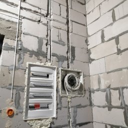 aWIĘC - ELEKTRO Patryk Więckiewicz Usługi Elektryczne - Rozdzielnica elektryczna z zabezpieczeniami, częściowo uszczelniona pianką montażową, zamontowana na surowej ścianie z betonu komórkowego, obok puszka z kablami.