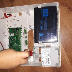 aWIĘC-ELEKTRO - Instalacja alarmowa w trakcie montażu: widoczna płyta główna, akumulator, transformator i dłoń dokręcająca śrubokrętem element wewnątrz białej obudowy.
