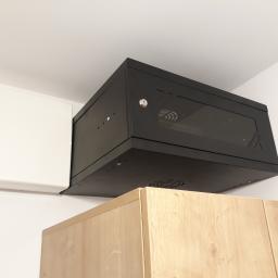 aWIĘC-ELEKTRO - Czarna szafka rackowa z przeszklonymi drzwiami zamontowana pod sufitem, obok biała skrzynka elektryczna, widoczny fragment drewnianej szafki pod spodem.