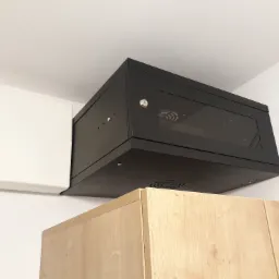 Czarna szafka rackowa z przeszklonymi drzwiami zamontowana pod sufitem, obok biała skrzynka elektryczna, widoczny fragment drewnianej szafki pod spodem.