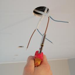 aWIĘC-ELEKTRO - Elektryk przy pracy: ręka trzyma ściągacz izolacji przy przewodach wychodzących z otworu w suficie.