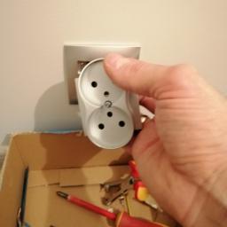 aWIĘC-ELEKTRO - Instalacja podwójnego gniazdka elektrycznego przez elektryka, widoczne narzędzia w kartonie poniżej, gniazdko w kolorze białym z srebrną ramką na ścianie.