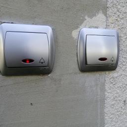 aWIĘC-ELEKTRO - Dwa srebrne przyciski dzwonkowe z czerwoną diodą, zamontowane na nierównej, szarej ścianie o zróżnicowanej fakturze.