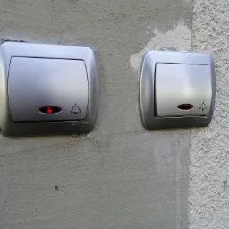 Dwa srebrne przyciski dzwonkowe z czerwoną diodą, zamontowane na nierównej, szarej ścianie o zróżnicowanej fakturze.