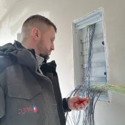 Elektryk w kurtce z logo firmy, podczas montażu przewodów w skrzynce rozdzielczej w surowym wnętrzu budynku.