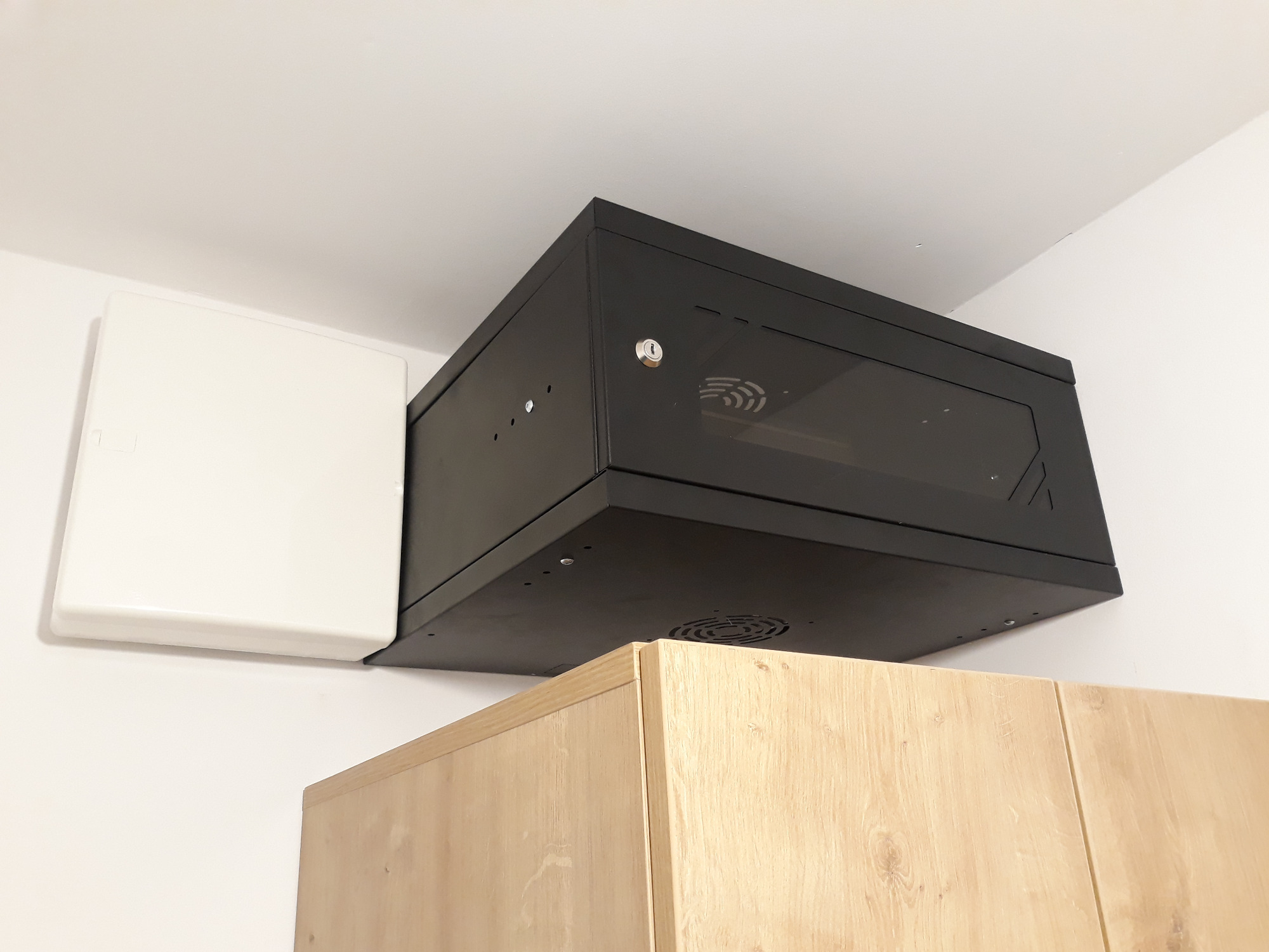 Czarna szafka rackowa z przeszklonymi drzwiami zamontowana pod sufitem, obok biała skrzynka elektryczna, widoczny fragment drewnianej szafki pod spodem.