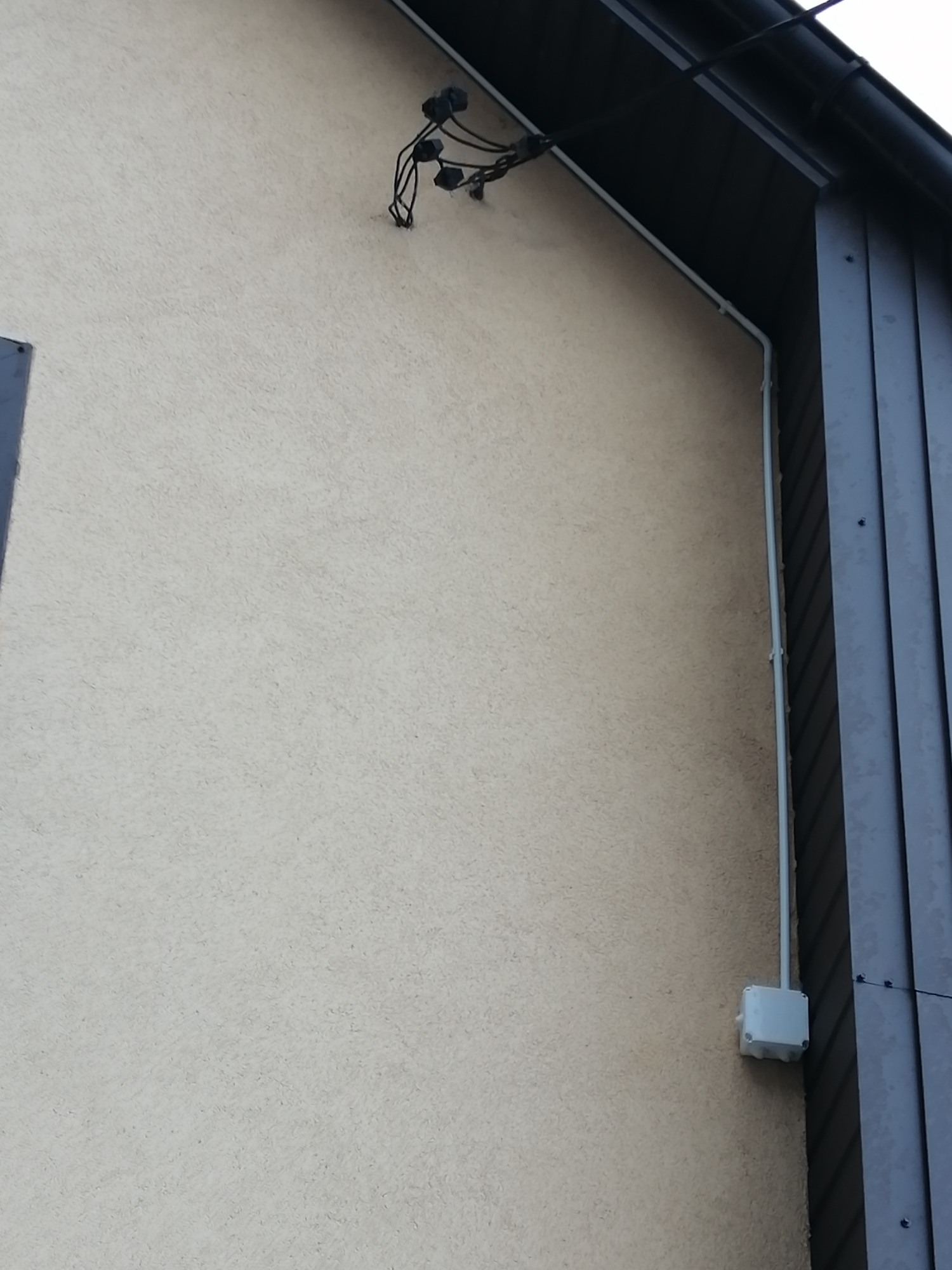 Biała elewacja budynku z widoczną instalacją elektryczną: kable wychodzące spod dachu, biała rurka z kablem biegnąca wzdłuż ściany oraz biała puszka elektryczna na dole.
