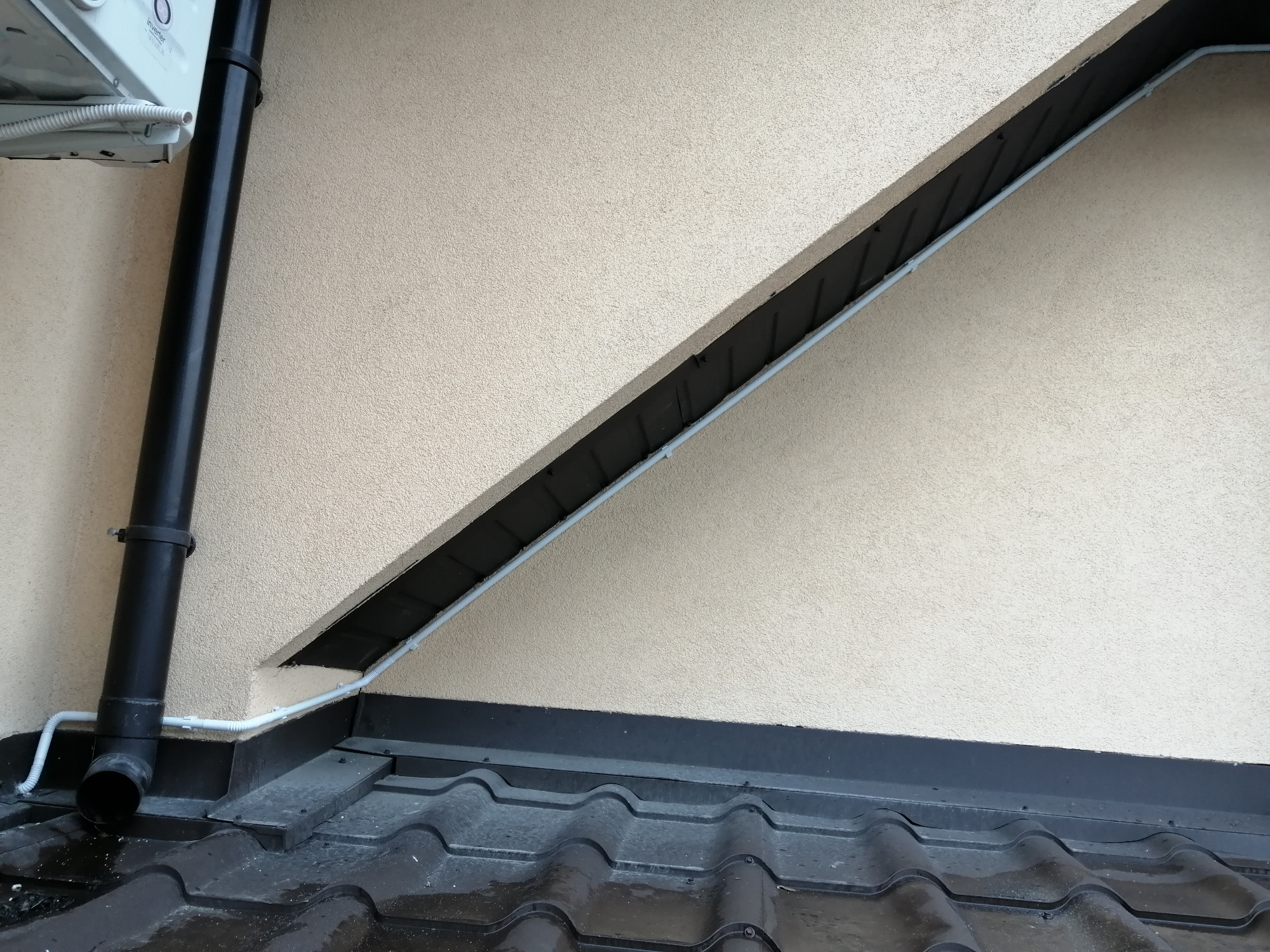 Biała rura karbowana prowadzona po elewacji budynku, od góry w dół, obok czarnej rury spustowej i dachu z blachodachówki.