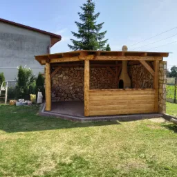 Drewniana altana z kamiennymi ścianami i piecem chlebowym w ogrodzie, z widoczną konstrukcją dachu i elementami wykończeniowymi.