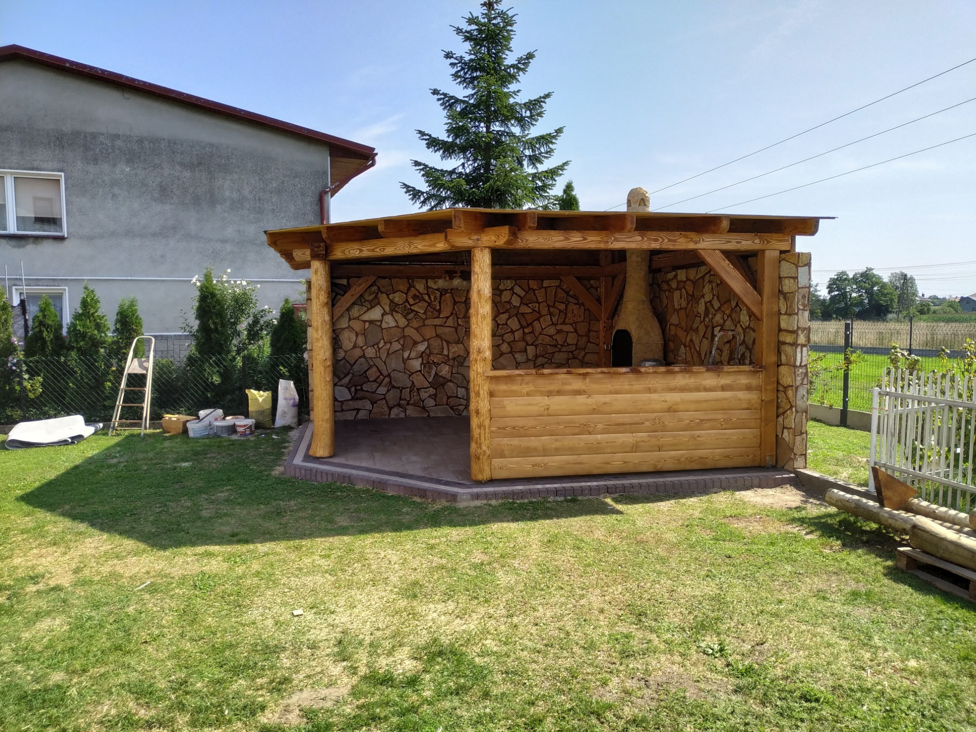 Drewniana altana z kamiennymi ścianami i piecem chlebowym w ogrodzie, z widoczną konstrukcją dachu i elementami wykończeniowymi.