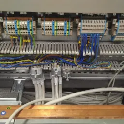 Wnętrze szafy elektrycznej z widocznymi listwami zaciskowymi, przewodami o różnych kolorach i oznaczeniach, oraz miedzianą szyną uziemiającą z naklejką 'N'.