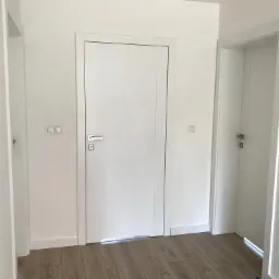 Białe, minimalistyczne drzwi wewnętrzne z srebrną klamką, zamontowane w białym korytarzu z panelami podłogowymi w odcieniach brązu, widoczne dwa inne wejścia.