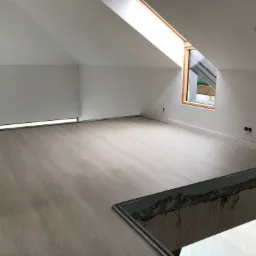 Poddasze z jasnymi panelami podłogowymi w trakcie montażu, widoczne paczki paneli przy ścianie, okno dachowe i niedokończone wykończenie schodów.