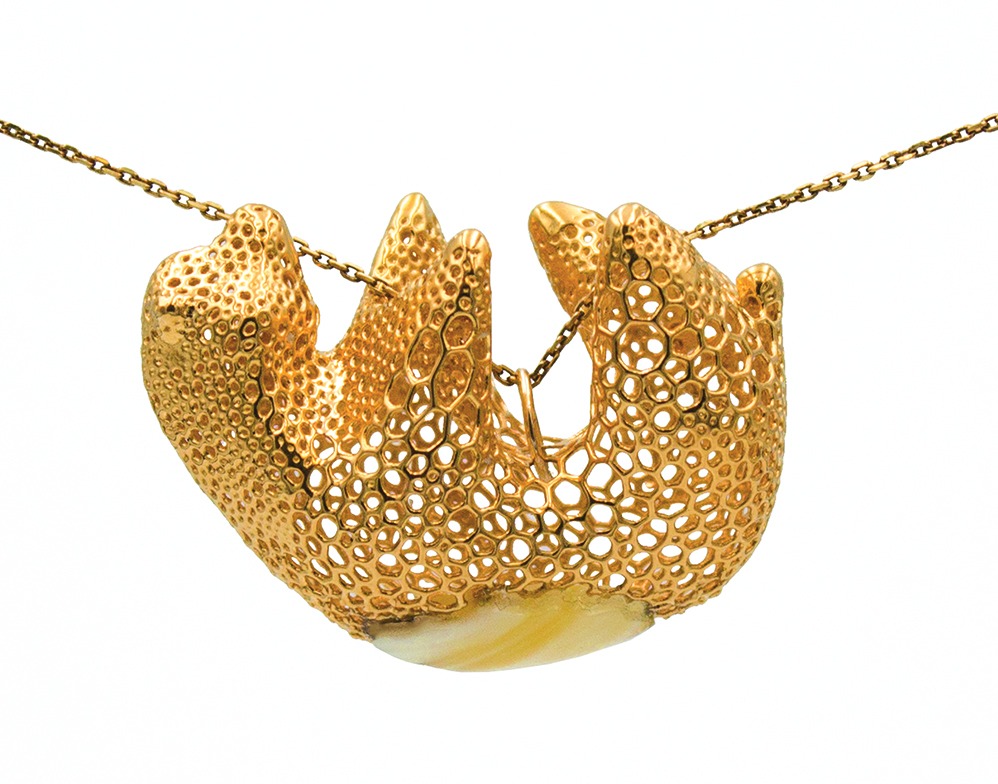Złoty wisiorek o ażurowej strukturze przypominającej gąbkę, zawieszony na delikatnym łańcuszku, z fragmentem bursztynu u dołu, sfotografowany na białym tle.