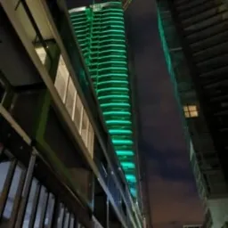 Nocny widok na nowoczesny, podświetlony na zielono wieżowiec w Koninie, widziany z dołu, z perspektywy ulicy, z widocznymi elementami konstrukcyjnymi i oknami sąsiednich budynków.