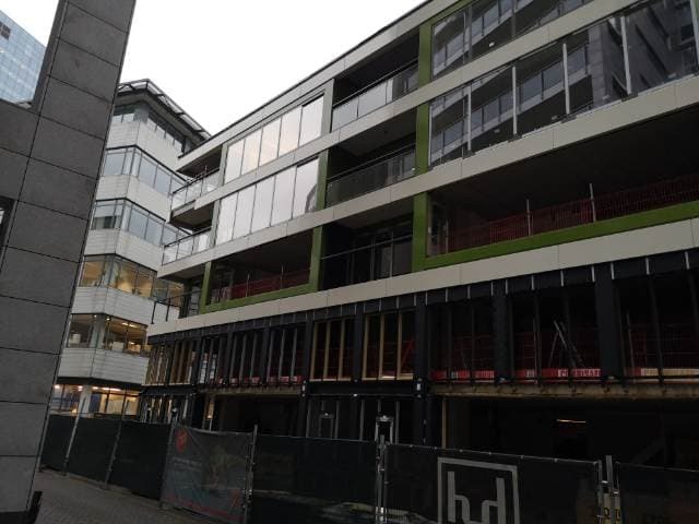 Wielopiętrowy budynek w trakcie wznoszenia elewacji, widoczne rusztowania i przygotowane ramy okienne na dolnych kondygnacjach, nowoczesna architektura w szarym otoczeniu miejskim.