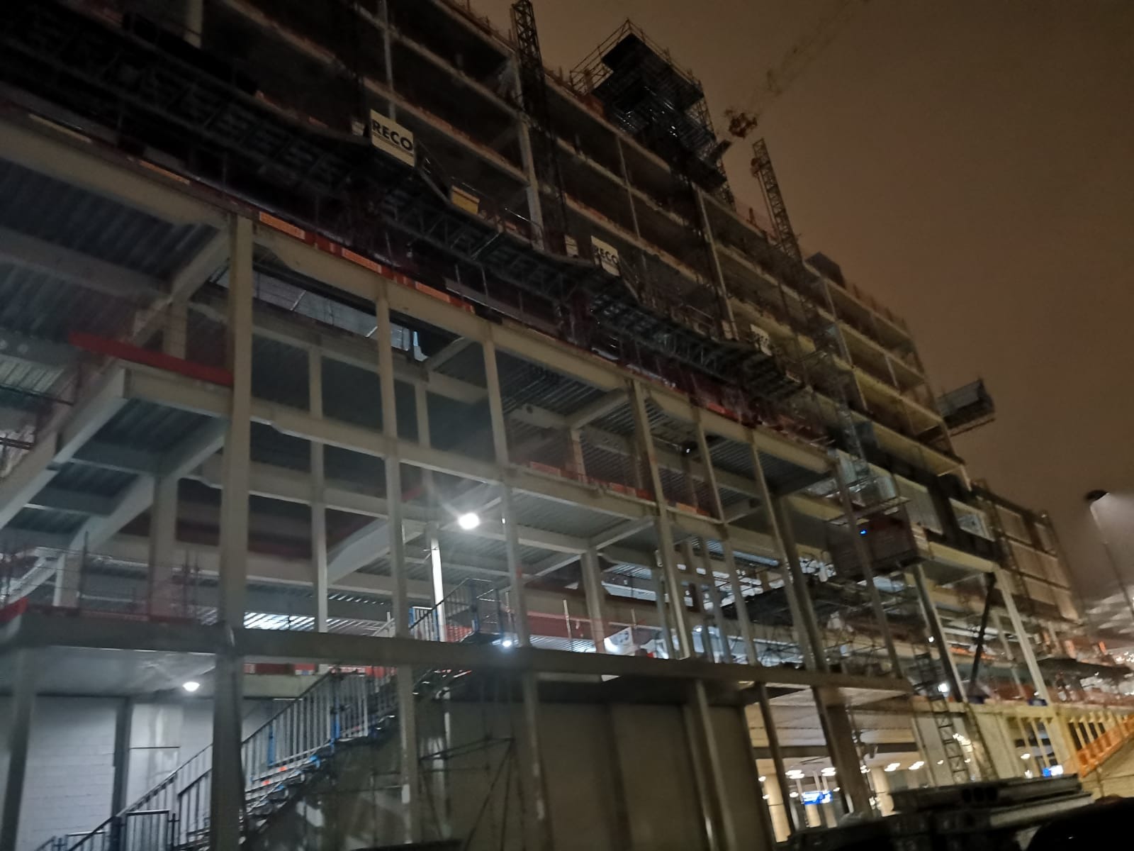 Nocne ujęcie konstrukcji szkieletowej budynku w trakcie budowy, widoczne stalowe belki i słupy, schody oraz żuraw budowlany w tle, oświetlone wewnętrznymi lampami.