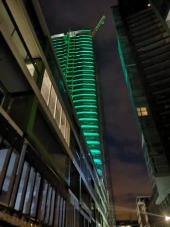 Nocny widok na nowoczesny, podświetlony na zielono wieżowiec w Koninie, widziany z dołu, z perspektywy ulicy, z widocznymi elementami konstrukcyjnymi i oknami sąsiednich budynków.