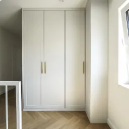 Biała szafa wnękowa z trzema skrzydłami i złotymi uchwytami, w jasnym wnętrzu z parkietem ułożonym w jodełkę. Widok z góry, z fragmentem białej balustrady.