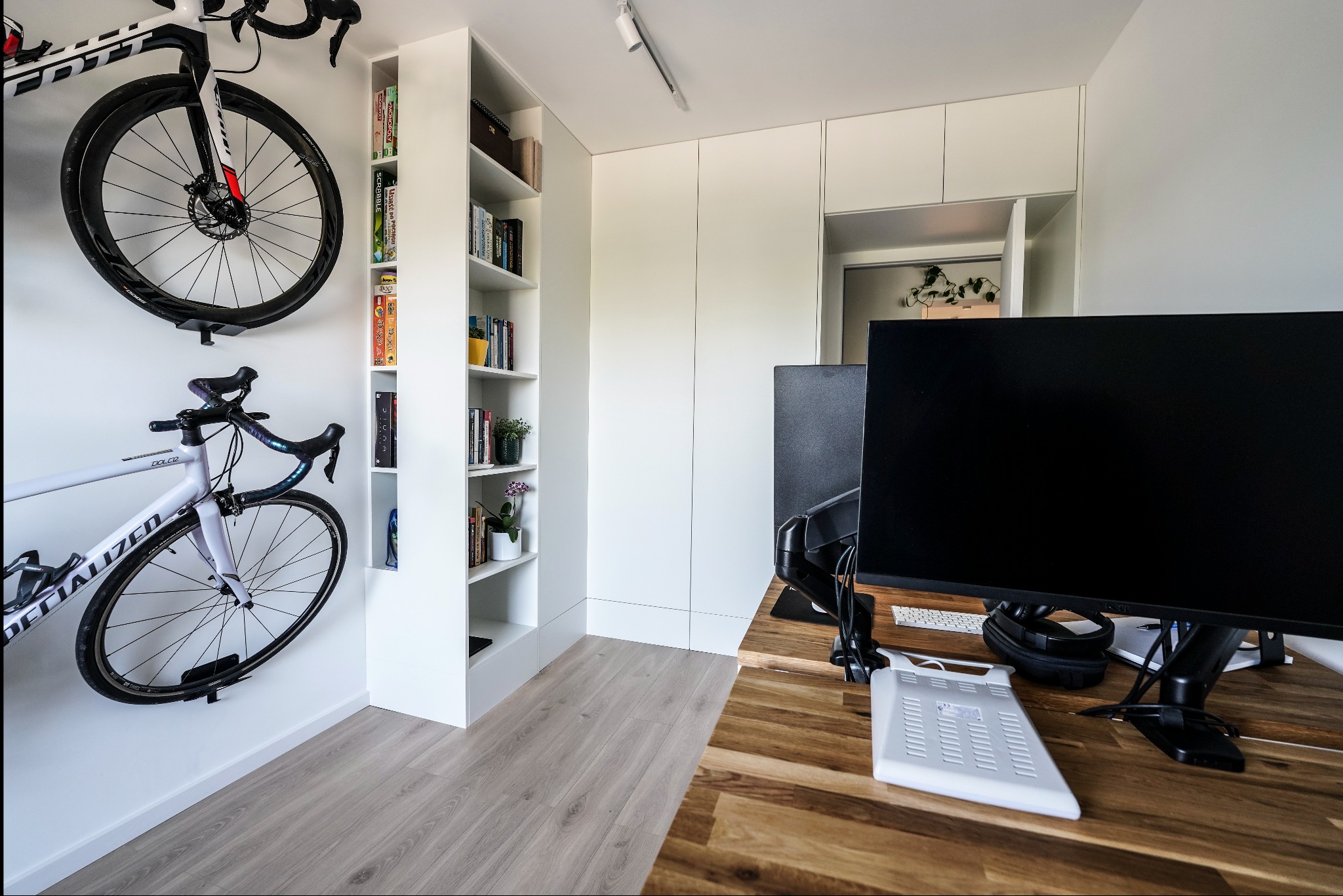 Minimalistyczne biuro z drewnianym blatem, białymi szafkami i regałem. Dwa rowery wiszą na ścianie, dodając sportowego charakteru. Nowoczesne oświetlenie.