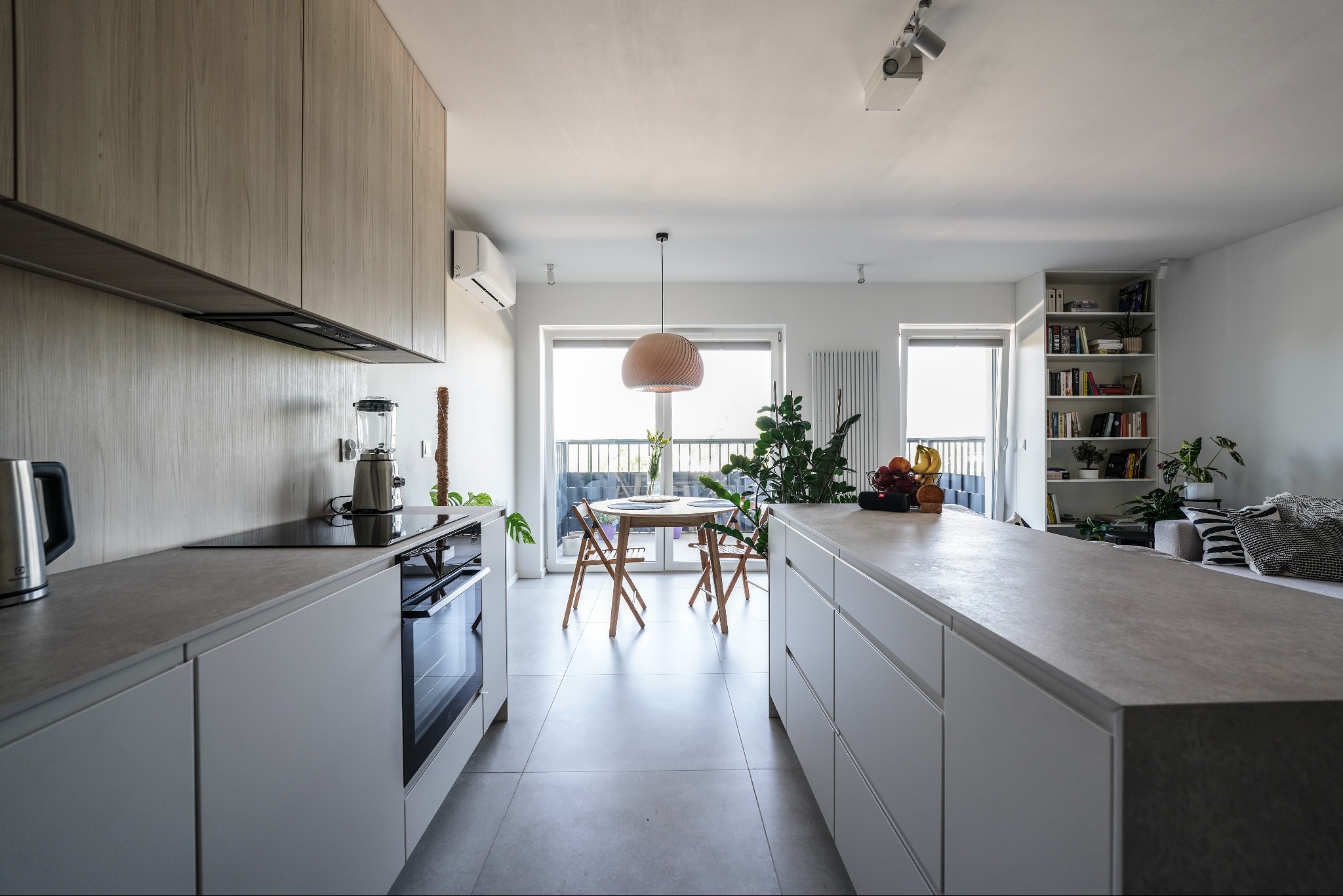 Jasna kuchnia z drewnianymi szafkami górnymi i białymi dolnymi, z wyspą i stołem jadalnym w tle. Minimalistyczny design, oświetlenie punktowe i naturalne światło.