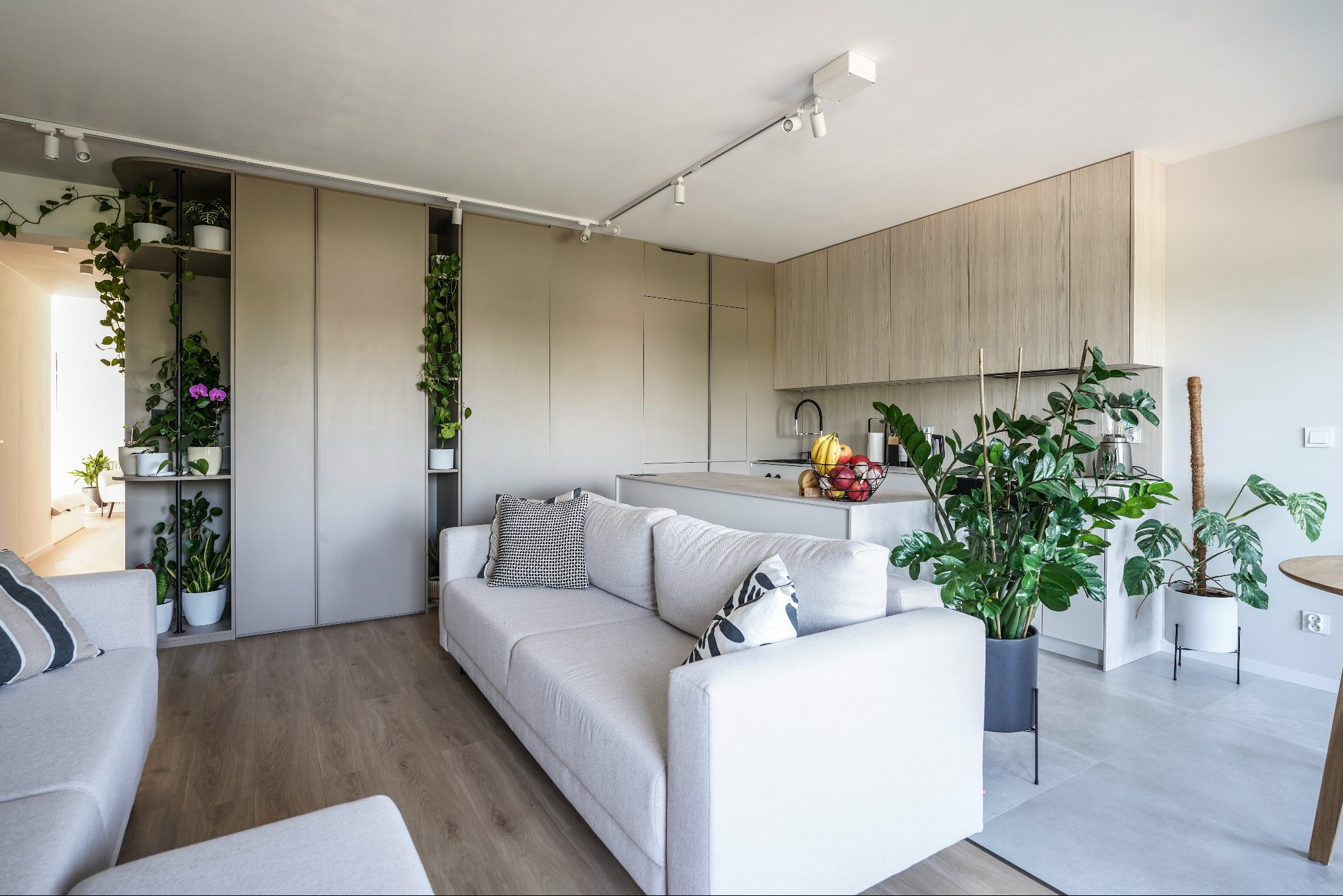 Jasne, nowoczesne wnętrze: kuchnia na wymiar z wyspą, minimalistyczne szafki, rośliny doniczkowe i sofa. Oświetlenie punktowe podkreśla detale.