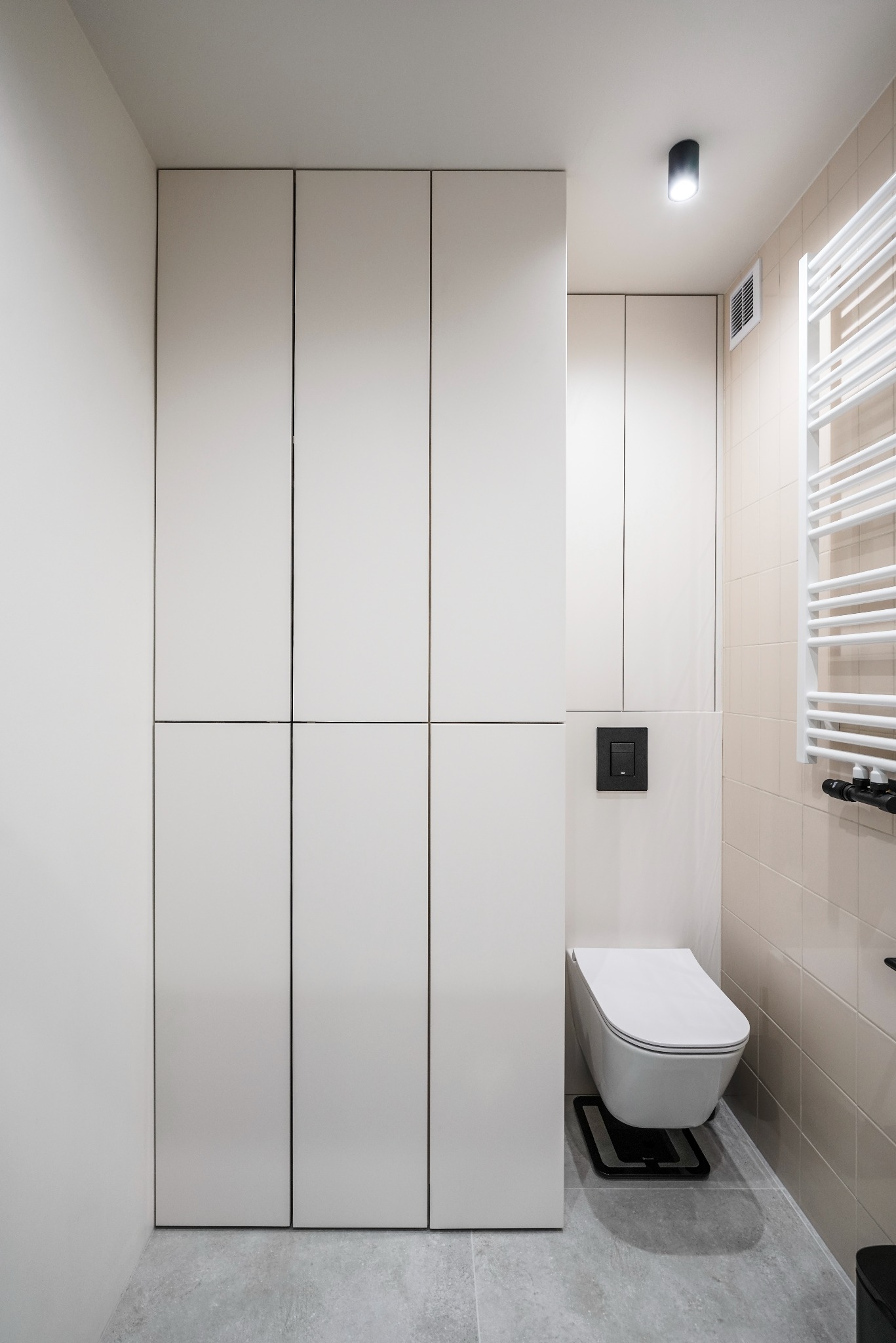 Jasna łazienka z zabudową meblową pod sufit, wiszącą toaletą i grzejnikiem drabinkowym na beżowych płytkach. Minimalistyczny design, szara podłoga.
