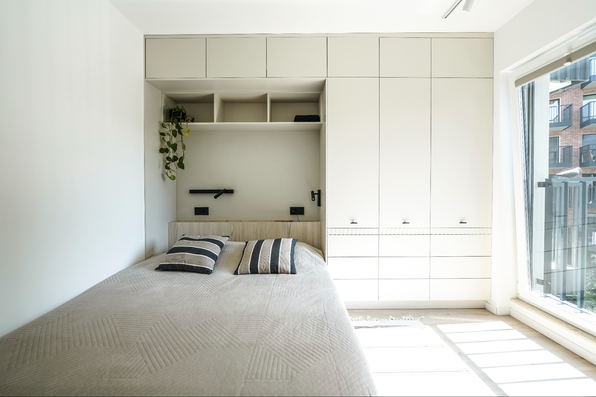 Nowoczesna sypialnia z zabudową meblową: białe szafy, półki, łóżko z wzorzystą narzutą, roślina pnąca i okno z widokiem na balkon.