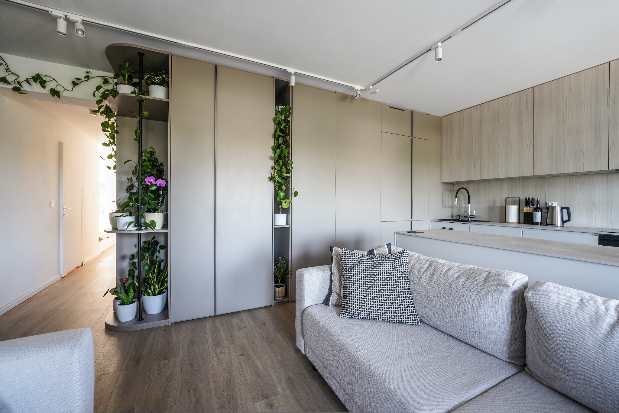 Nowoczesna kuchnia z zabudową w odcieniach szarości, z roślinami wkomponowanymi w regały. Widoczny fragment salonu z sofą i drewnianą podłogą.