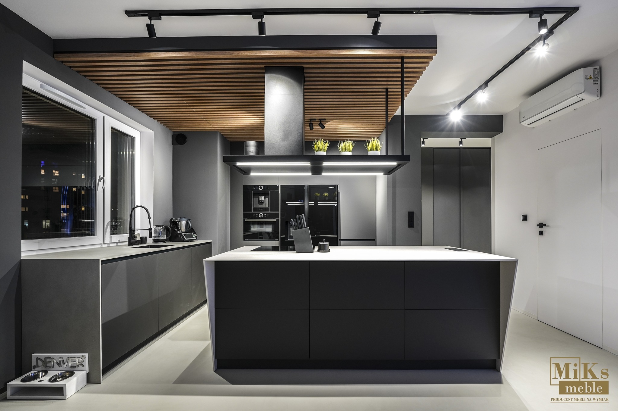 Nowoczesna, szara kuchnia z wyspą i drewnianą podsufitką. Minimalistyczny design, czarne sprzęty AGD, oświetlenie punktowe. Widok z przodu, logo w prawym dolnym rogu.