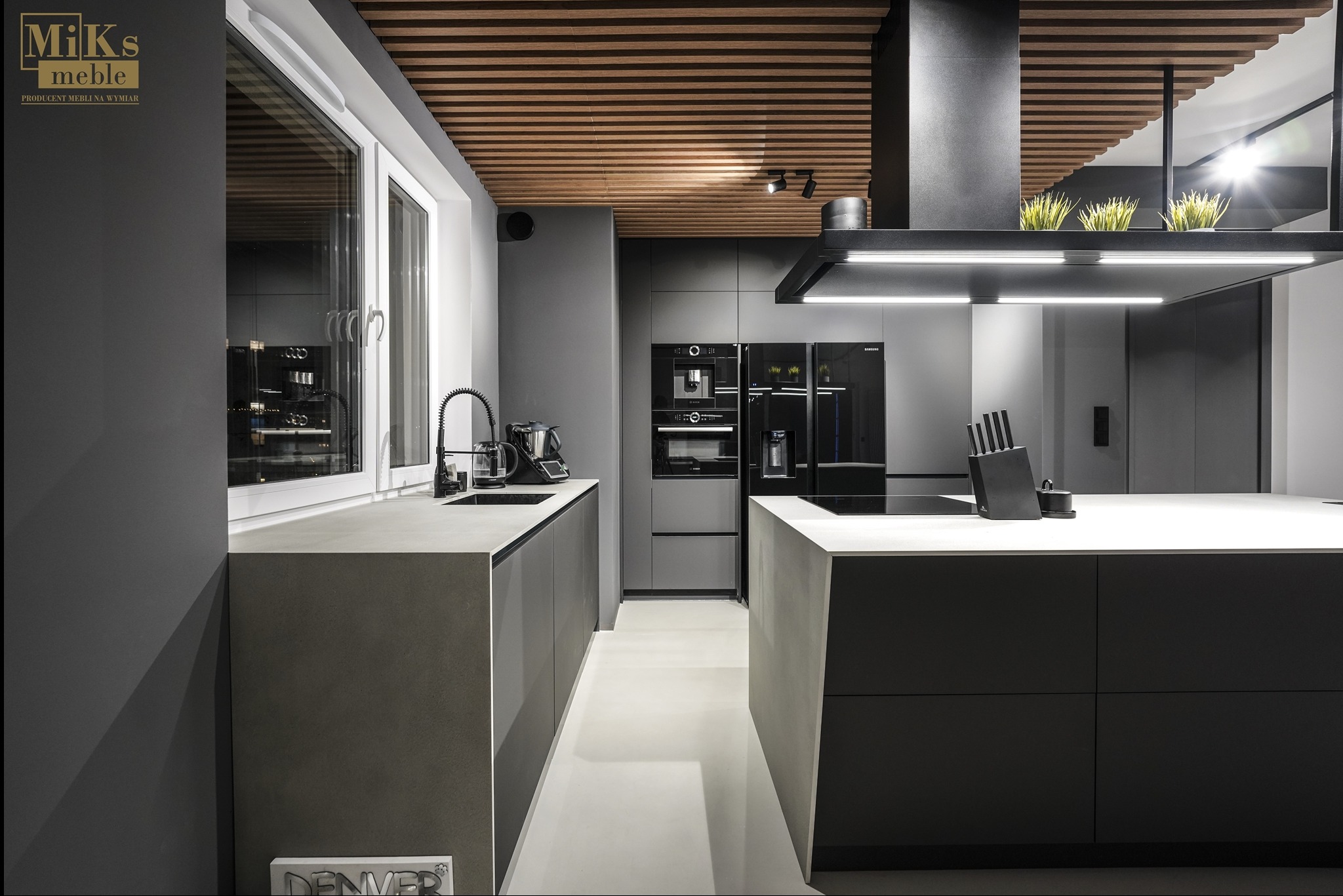 Nowoczesna, minimalistyczna kuchnia w odcieniach szarości z drewnianymi elementami na suficie i wyspą kuchenną. Widoczne sprzęty AGD i akcesoria.