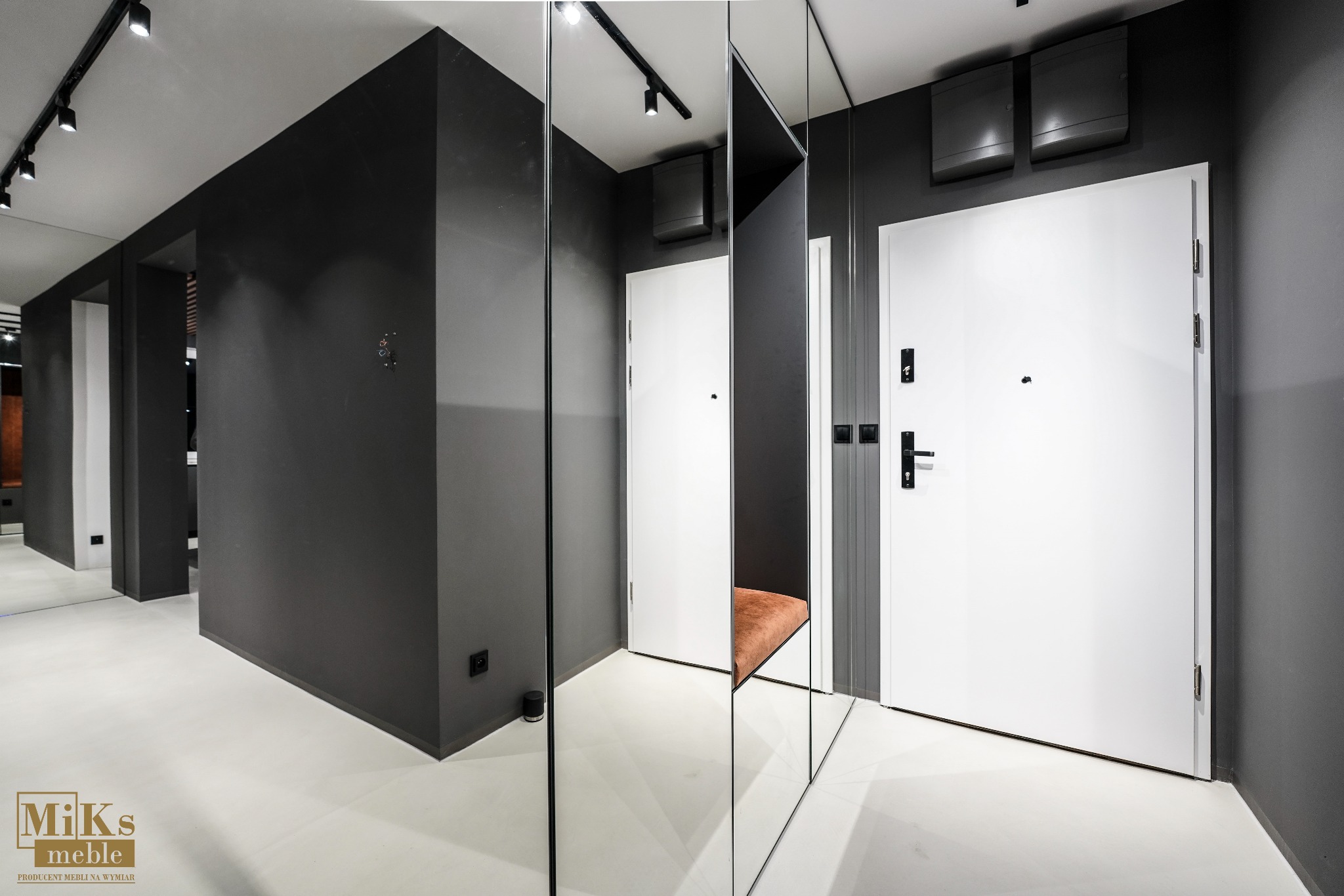 Nowoczesny przedpokój w odcieniach szarości z lustrami i białymi drzwiami. Widoczne oświetlenie punktowe i minimalistyczne wykończenie. Na podłodze widoczne logo producenta mebli.
