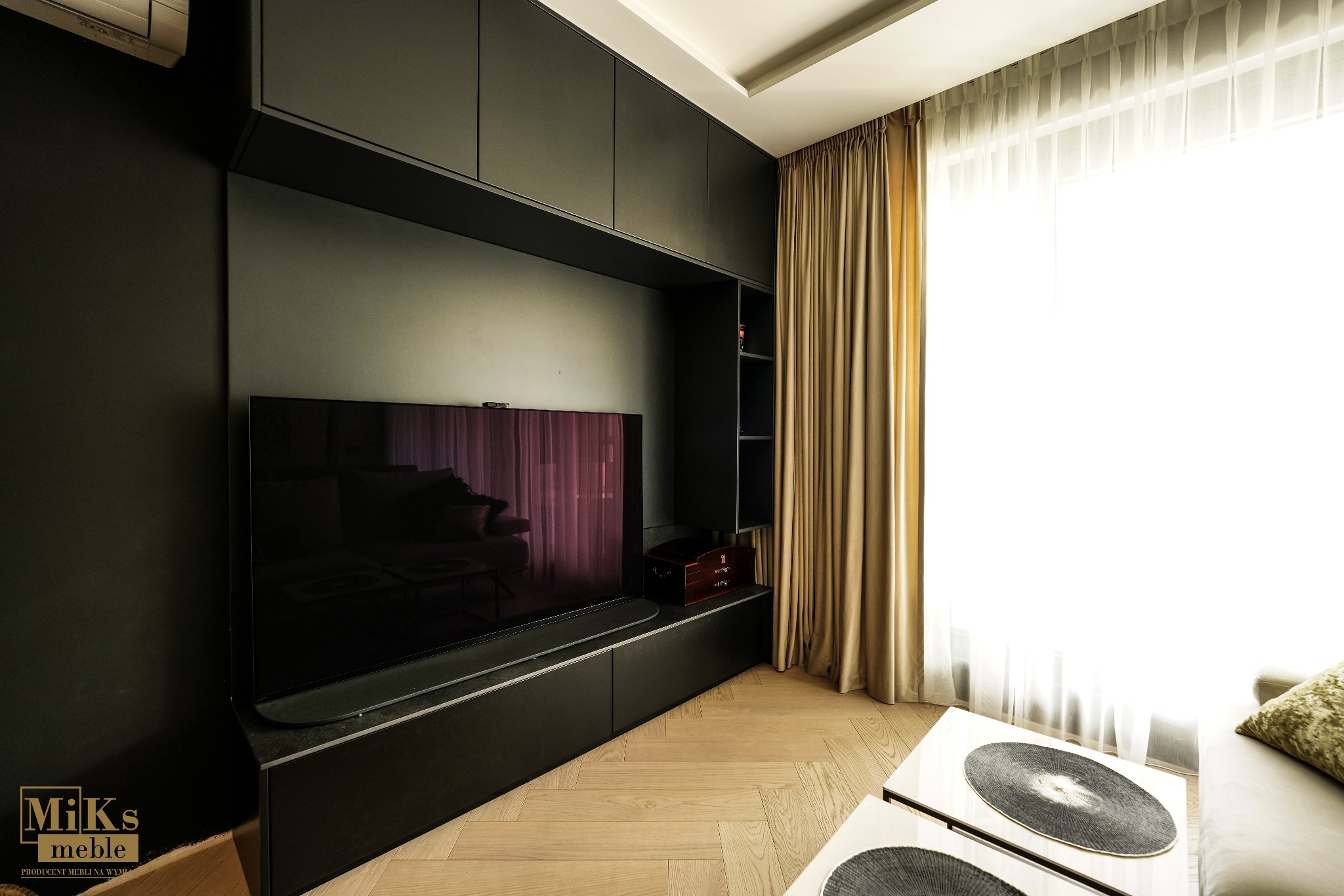 Nowoczesny salon z zabudową meblową na ścianie w odcieniach szarości, pod telewizor. Parkiet ułożony w jodełkę. Długie zasłony w oknie.