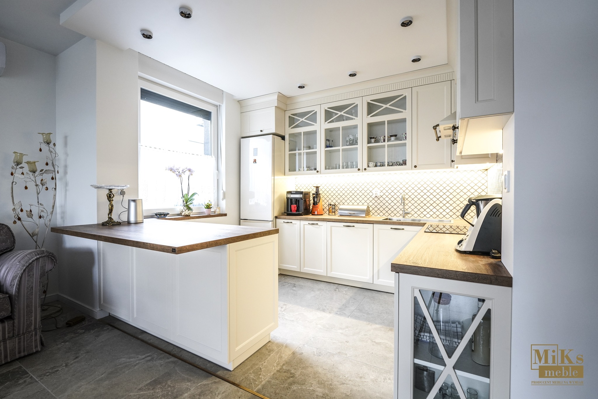 Jasna kuchnia z wyspą i drewnianymi blatami, przeszklone szafki wiszące, mozaika na ścianie, nowoczesny design, marmurowa podłoga i dekoracje.