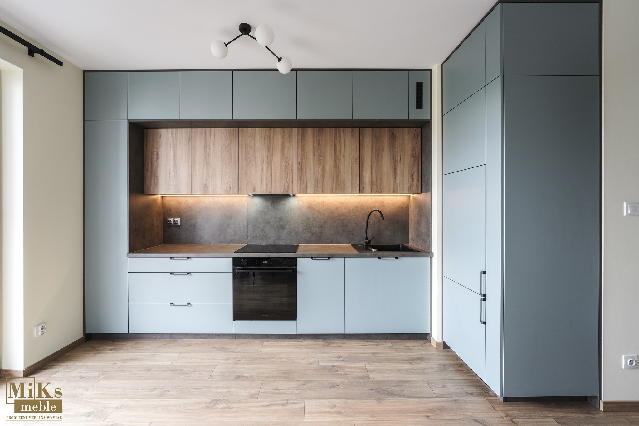 Nowoczesna kuchnia: błękitne szafki, drewniane górne półki i szary blat z oświetleniem LED. Minimalistyczna lampa sufitowa i drewniana podłoga dodają elegancji.
