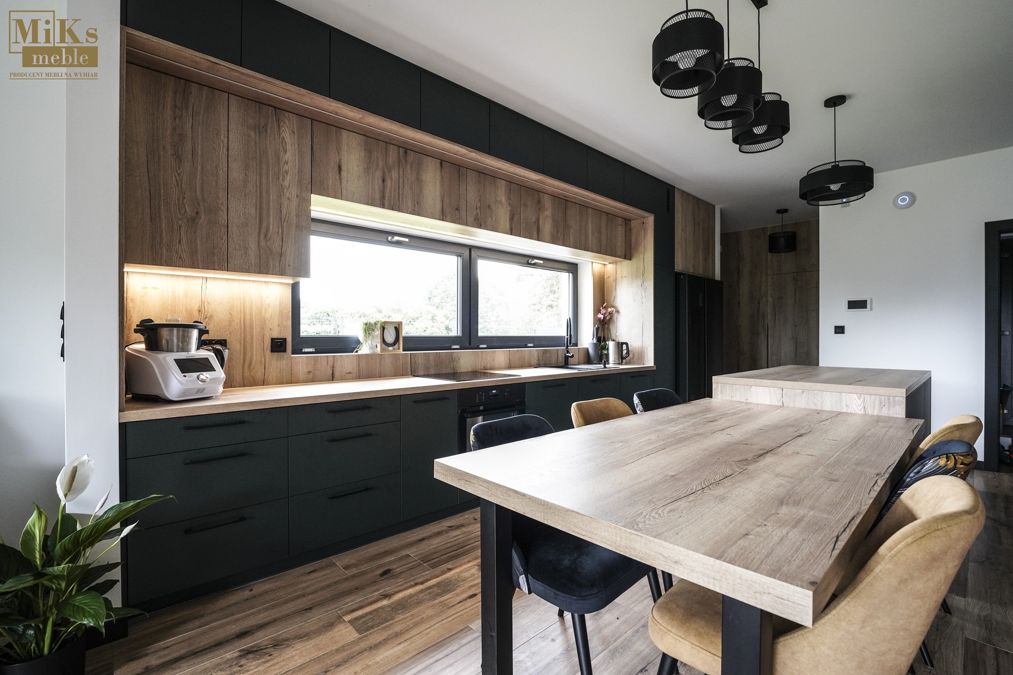 Nowoczesna kuchnia z ciemnozielonymi szafkami i drewnianymi blatami. Duże okno nad blatem roboczym. Stół z krzesłami w centrum, lampy wiszące. Parkiet na podłodze.