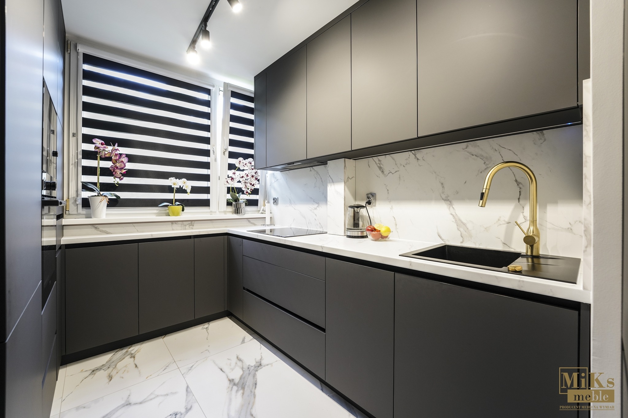Nowoczesna kuchnia na wymiar z szarymi frontami, białym blatem i marmurowym backsplash. Złota bateria, czarny zlew i rolety w oknie dodają elegancji.